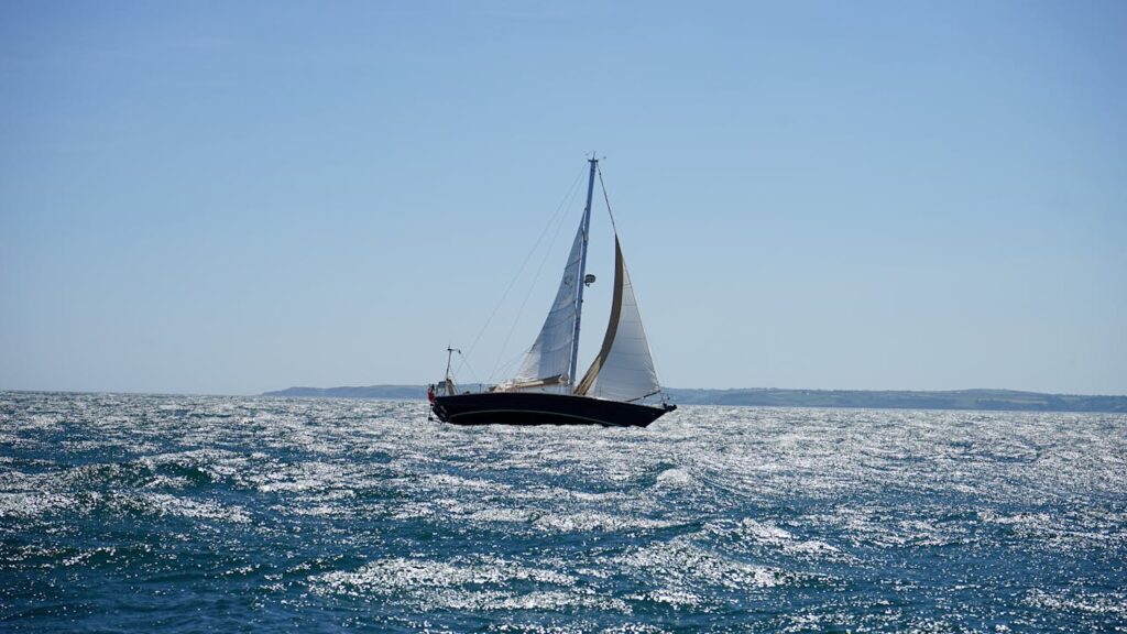 A sailboat gracefully navigates the sparkling ocean off the coast of Cornwall on a sunny summer day.